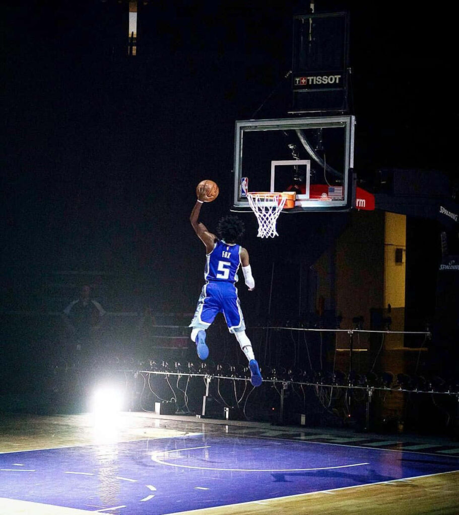 De'Aaron Fox mid-air dunk in blue white uniform against dark arena high energy basketball scene Wallpaper