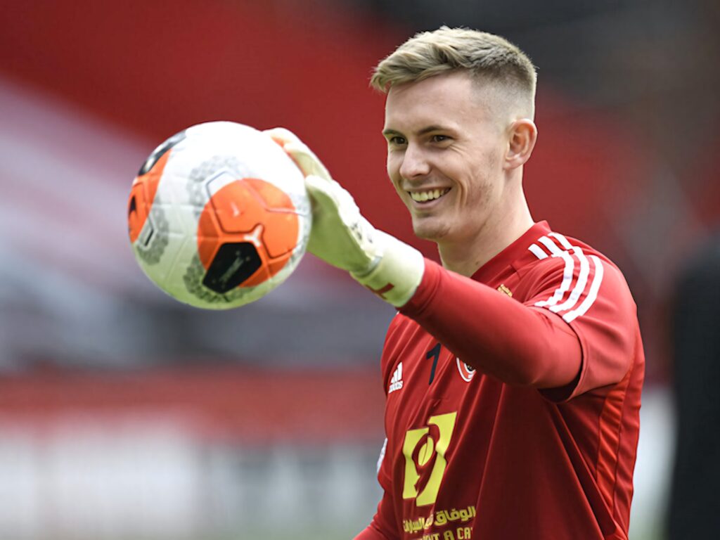 Dean Henderson in red goalkeeper kit smiling confidently holding soccer ball stadium background wallpaper