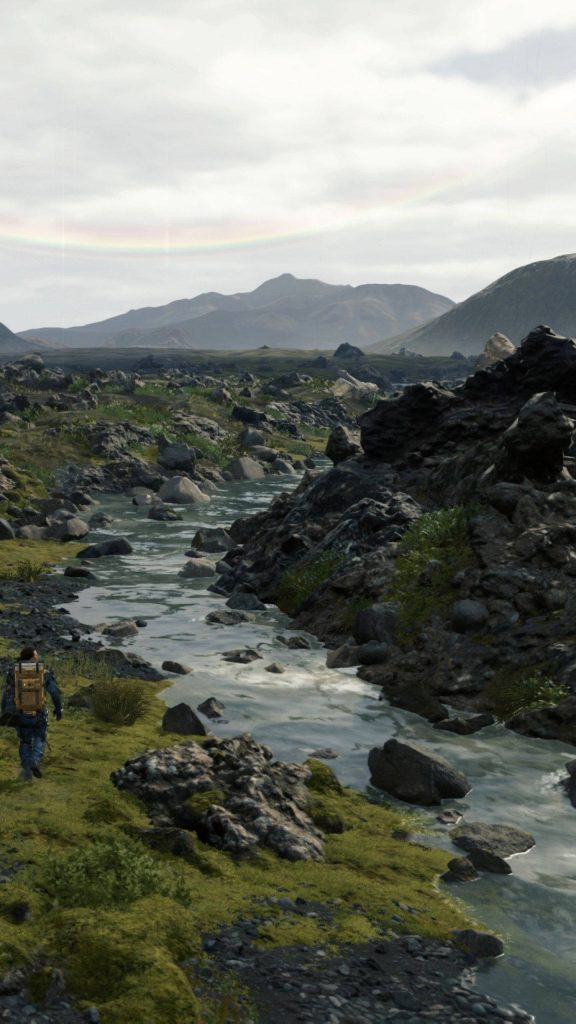 Sam Porter Bridges trekking rugged mossy terrain beside stream with rainbow Death Stranding wallpaper