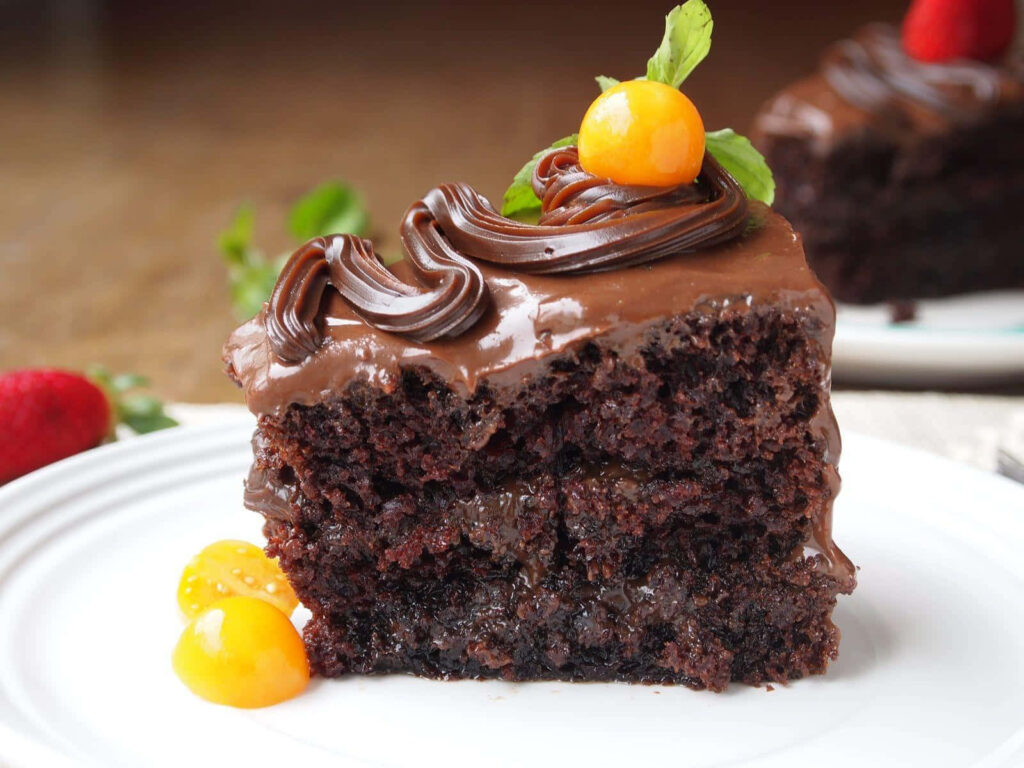 Slice of rich chocolate cake with glossy frosting golden berries and fresh mint on white plate Wallpaper