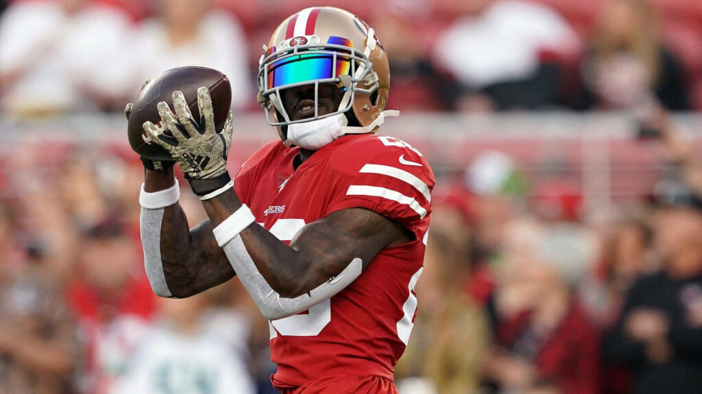 Deebo Samuel Sr throwing football in red and gold 49ers uniform with vibrant stadium background Wallpaper