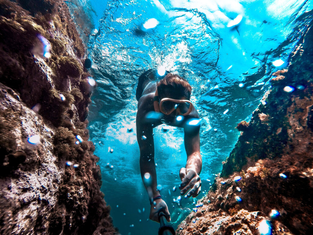 Deep sea diver swimming through rocky formations in clear blue water with sunlight and bubbles Wallpaper