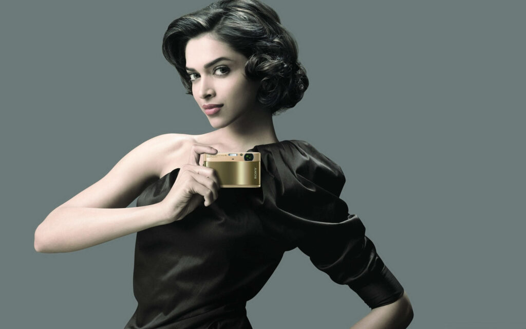 Deepika Padukone in black dress holding gold Sony camera against minimalist grey background Wallpaper