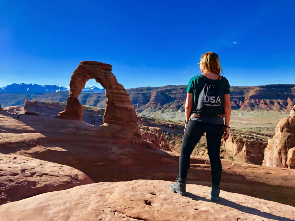 Delicate Arch landscape with red rocks blue sky and hiker carrying USA backpack in desert vista Wallpaper