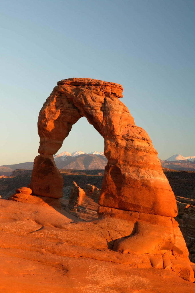 Delicate Arch glowing in sunlight with red sandstone and snow-capped mountains in Arches Park Wallpaper