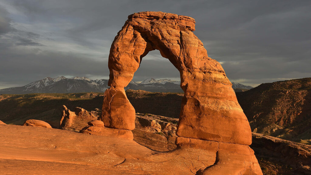 Delicate Arch glowing red sandstone under dark skies with distant mountains desert landscape wallpaper