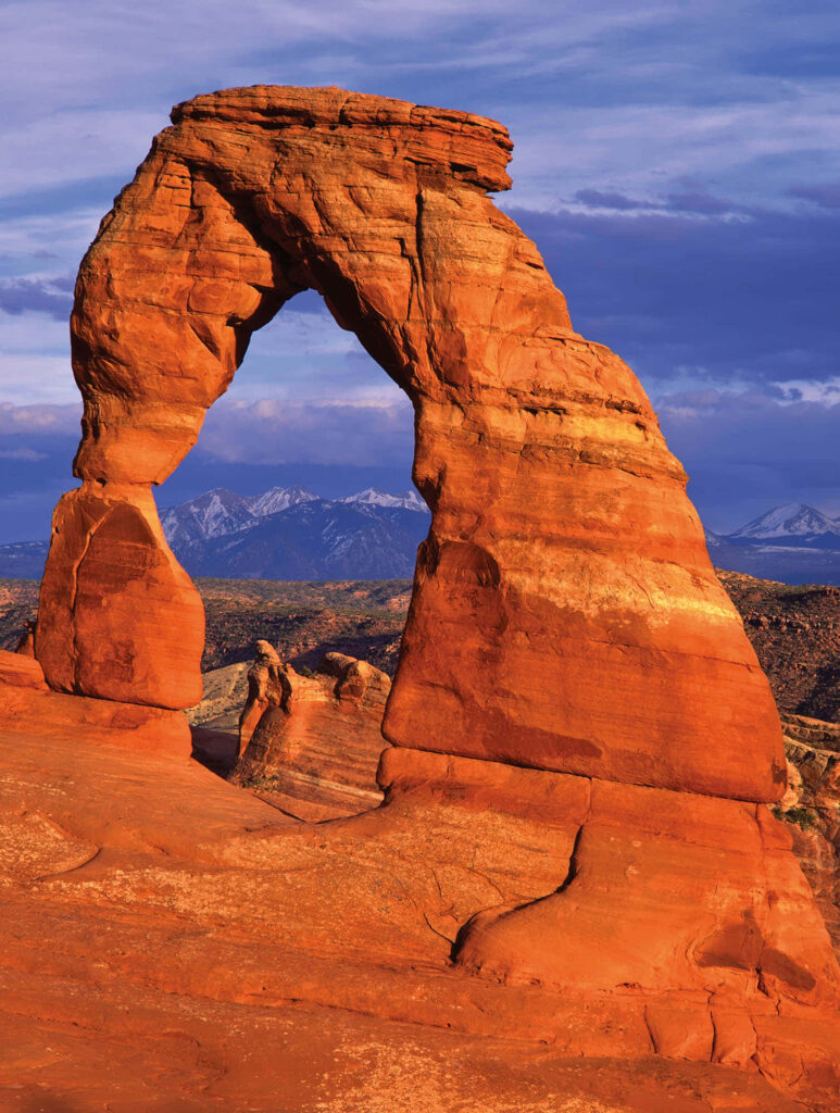 Delicate Arch glowing orange red at sunset with smooth wind-carved edges and mountain backdrop Wallpaper