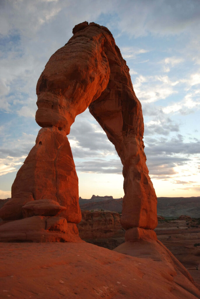 Delicate Arch glowing red orange at sunset against soft clouds in rugged desert landscape wallpaper