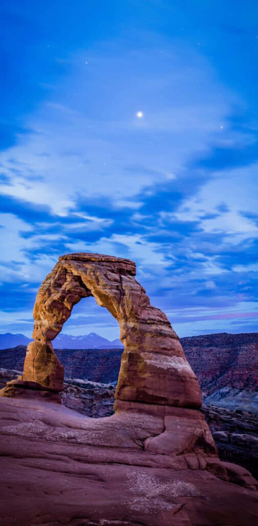 Delicate Arch at twilight with red sandstone glowing under soft blue clouds and shining stars Wallpaper