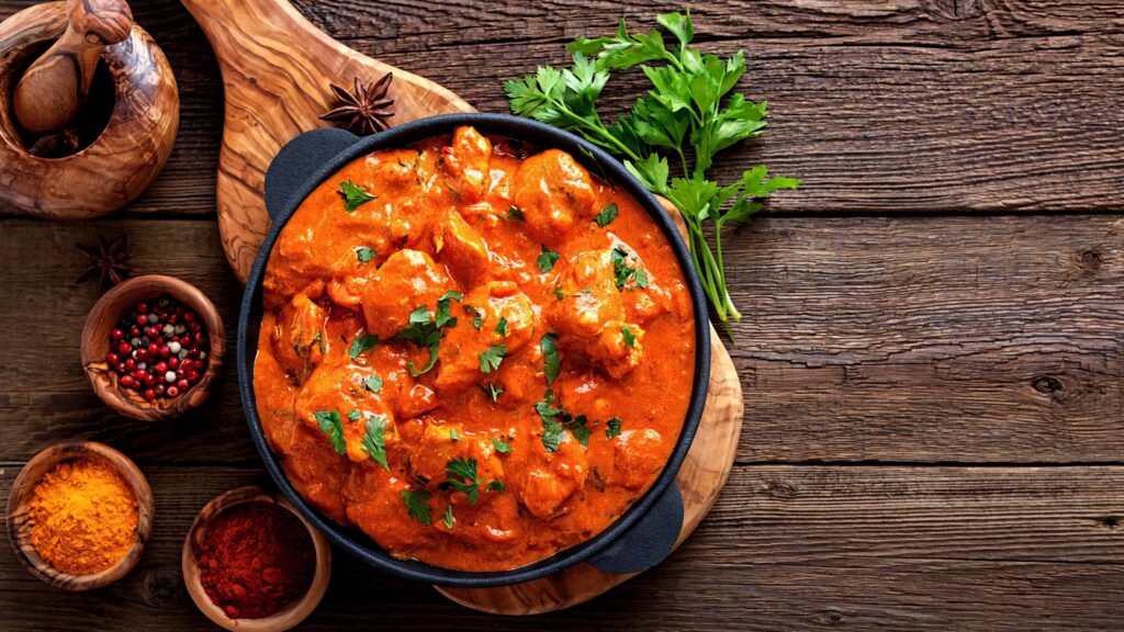Butter chicken in dark bowl with fresh herbs and spices on rustic wooden board wallpaper background
