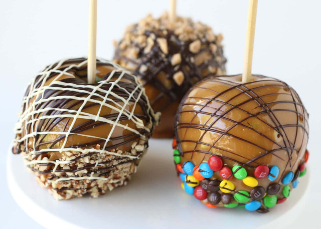 Three caramel apples with nuts candies and chocolate drizzle on white festive background wallpaper