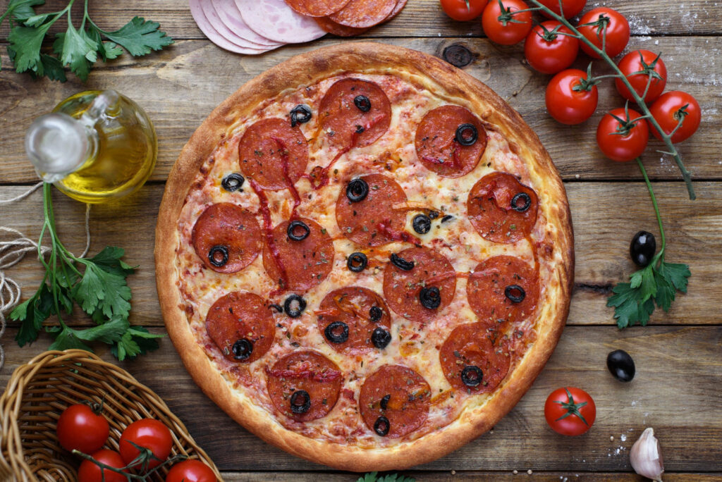 Large pepperoni pizza with black olives on rustic table surrounded by fresh ingredients and herbs Wallpaper