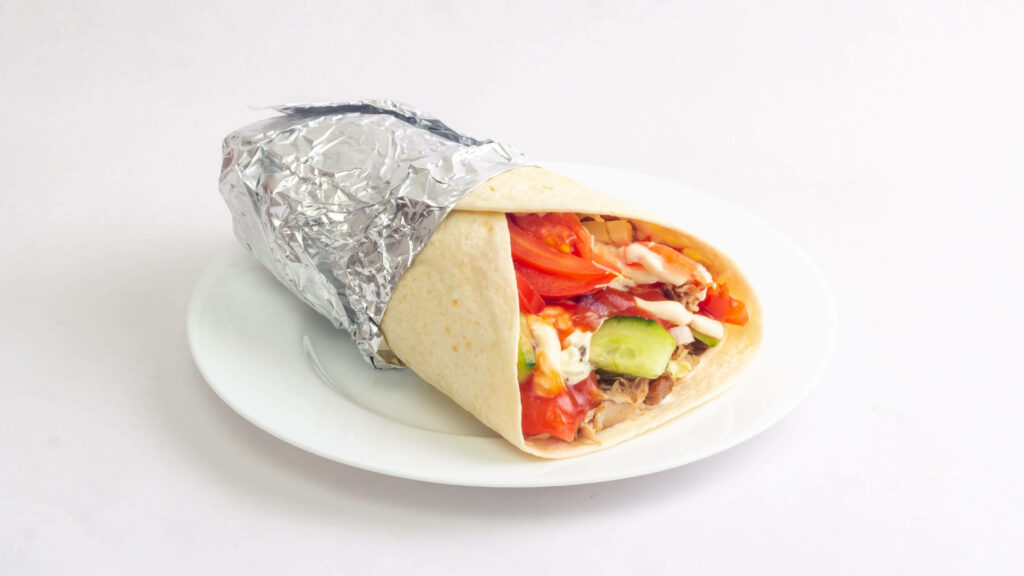 Shawarma burrito wrapped in foil on white plate with fresh tomatoes lettuce and sauce background Wallpaper
