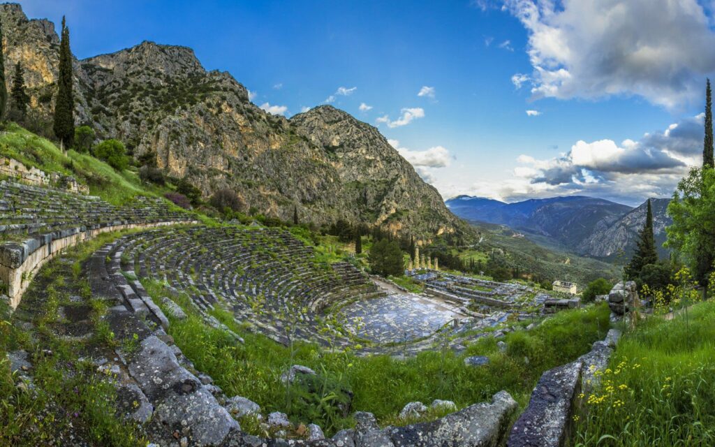 Ancient Delphi ruins stone amphitheater with green grass wildflowers mountains blue sky wallpaper