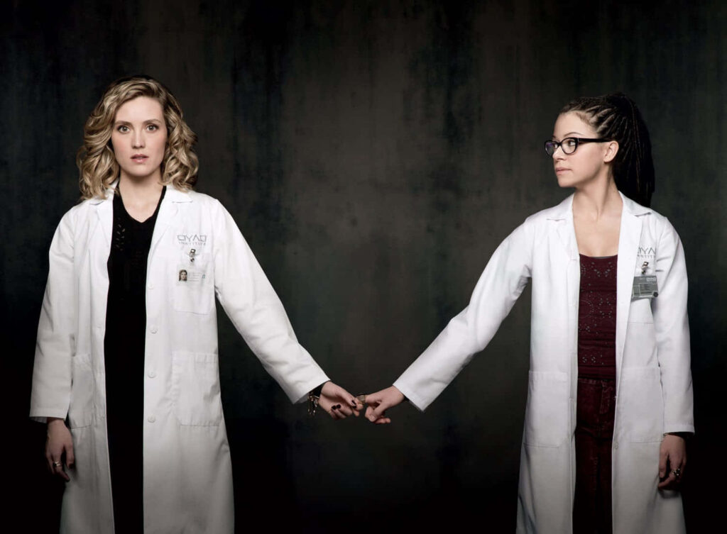 Delphine and Cosima from Orphan Black in lab coats holding hands against dark background wallpaper