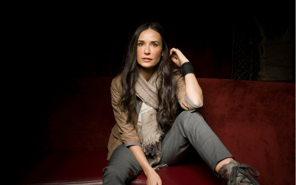 Demi Moore seated on red sofa in casual outfit with brown jacket and textured scarf portrait Wallpaper