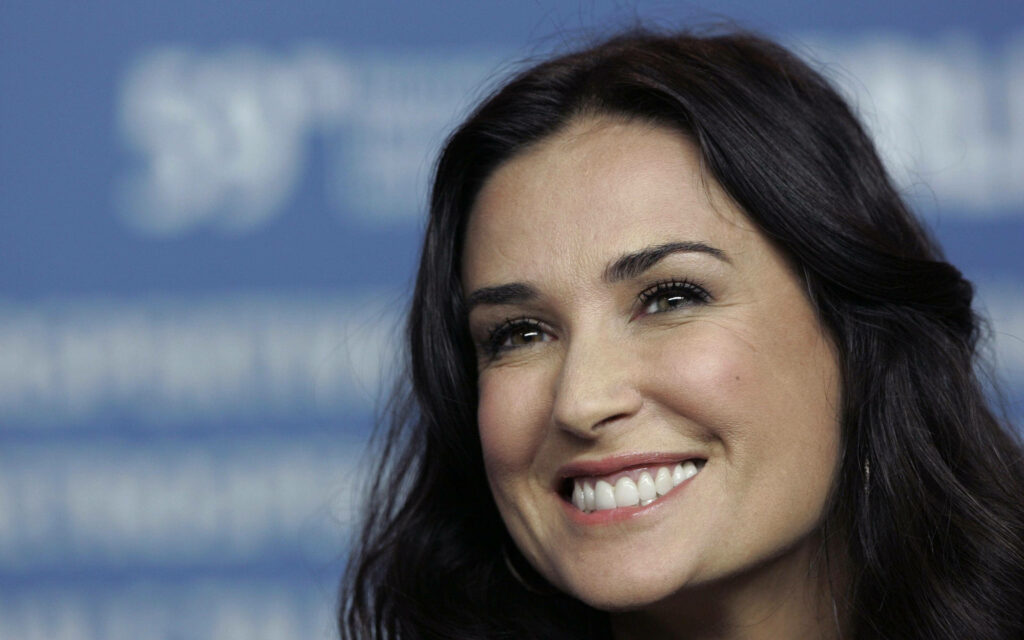 Demi Moore close-up portrait with radiant smile glowing complexion dark flowing hair soft blue background Wallpaper