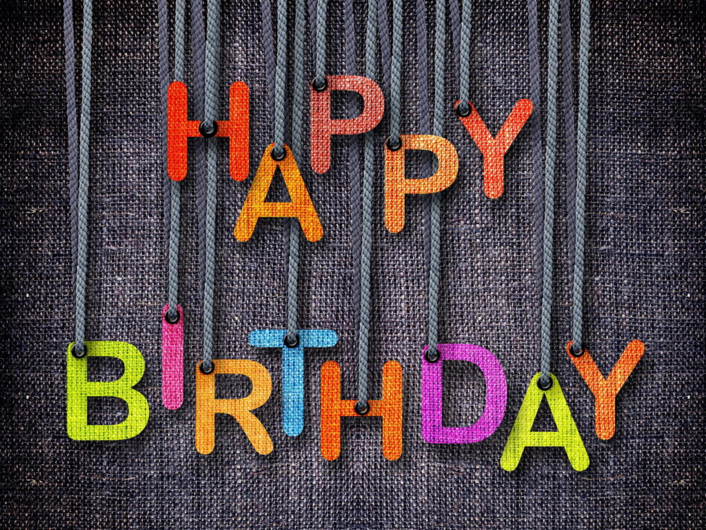 Happy Birthday multicolored fabric letters hanging on denim ropes dark woven background wallpaper
