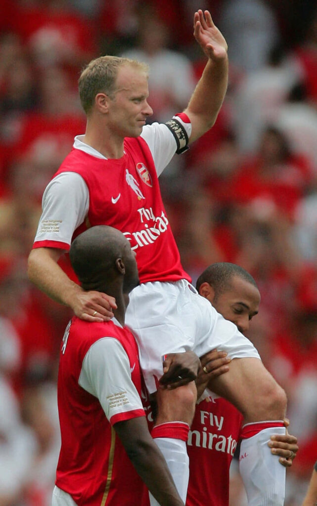 Dennis Bergkamp lifted by Arsenal teammates in red and white kits during packed stadium celebration Wallpaper