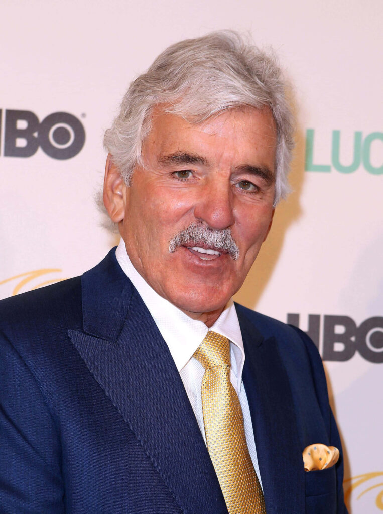 Dennis Farina in navy suit and yellow tie posing confidently at bright HBO event background wallpaper