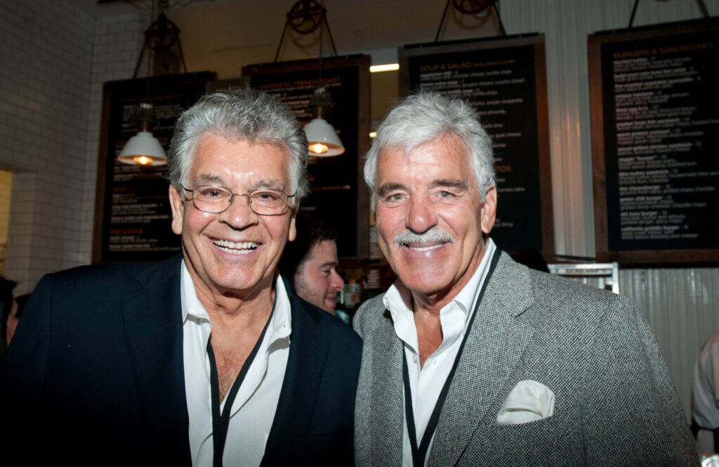 Dennis Farina and Kevin Riley in elegant suits smiling in upscale restaurant with warm lighting Wallpaper