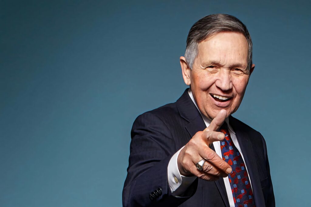 Dennis Kucinich in dark suit gestures confidently against blue gradient background portrait wallpaper