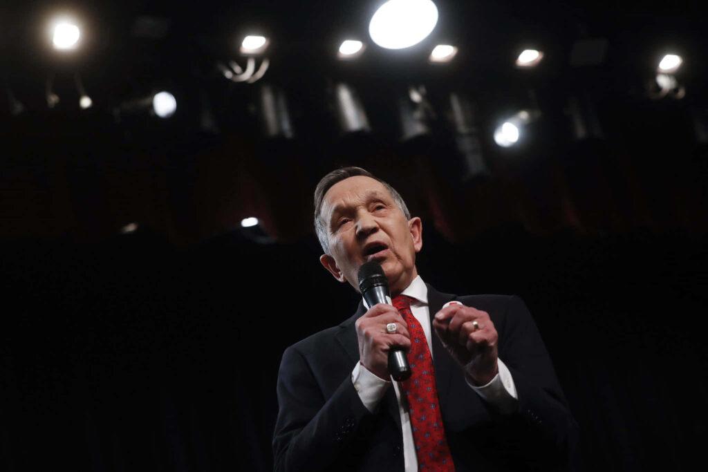 Dennis Kucinich speaking passionately in dark suit red tie dramatic stage lighting black backdrop Wallpaper