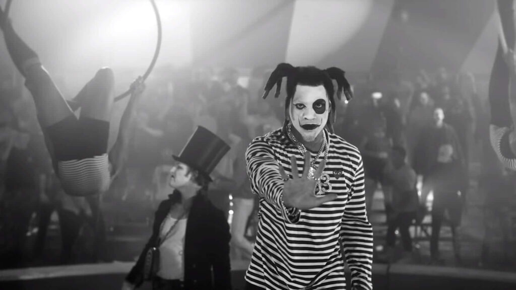 Denzel Curry as dramatic clown in striped outfit with bold face paint at lively circus event background Wallpaper