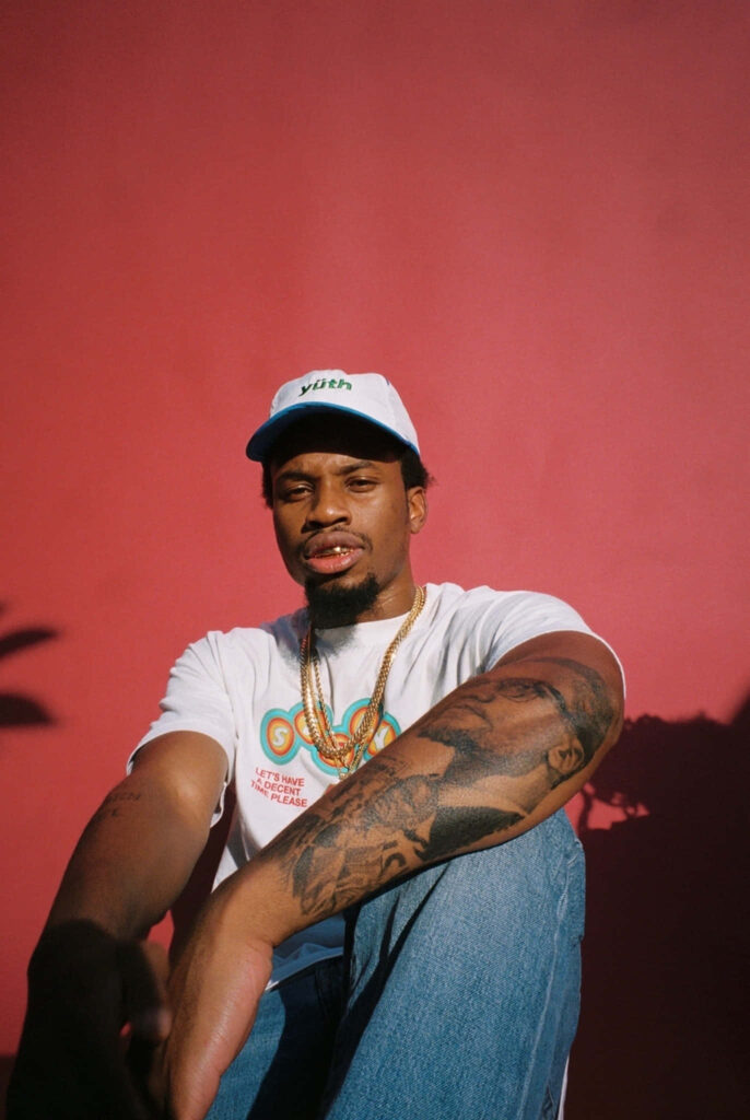 Denzel Curry in white tee and baseball cap against red background featuring gold accessories tattoo Wallpaper