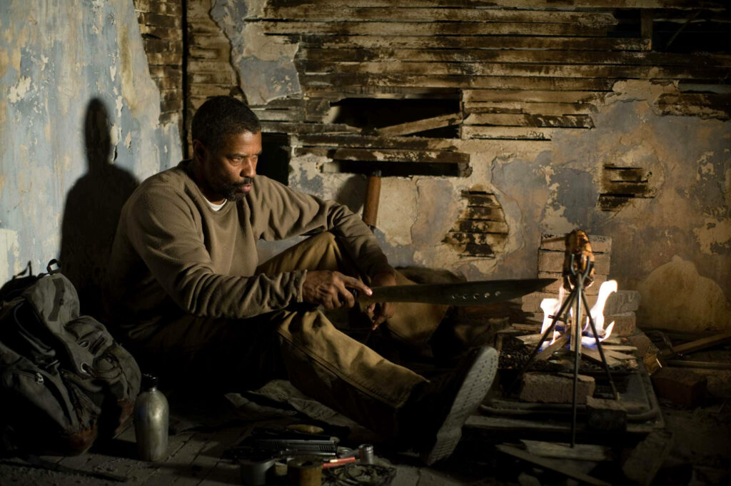Denzel Washington sharpening a large machete in a dim rundown room by fire intense gritty scene Wallpaper