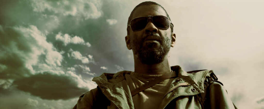 Denzel Washington in sunglasses and rugged attire stands confidently under dramatic cloudy sky background Wallpaper