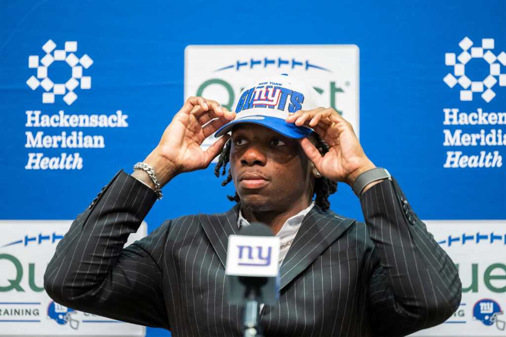 Deonte Banks in pinstripe suit adjusting blue NY Giants cap at official team signing event backdrop Wallpaper