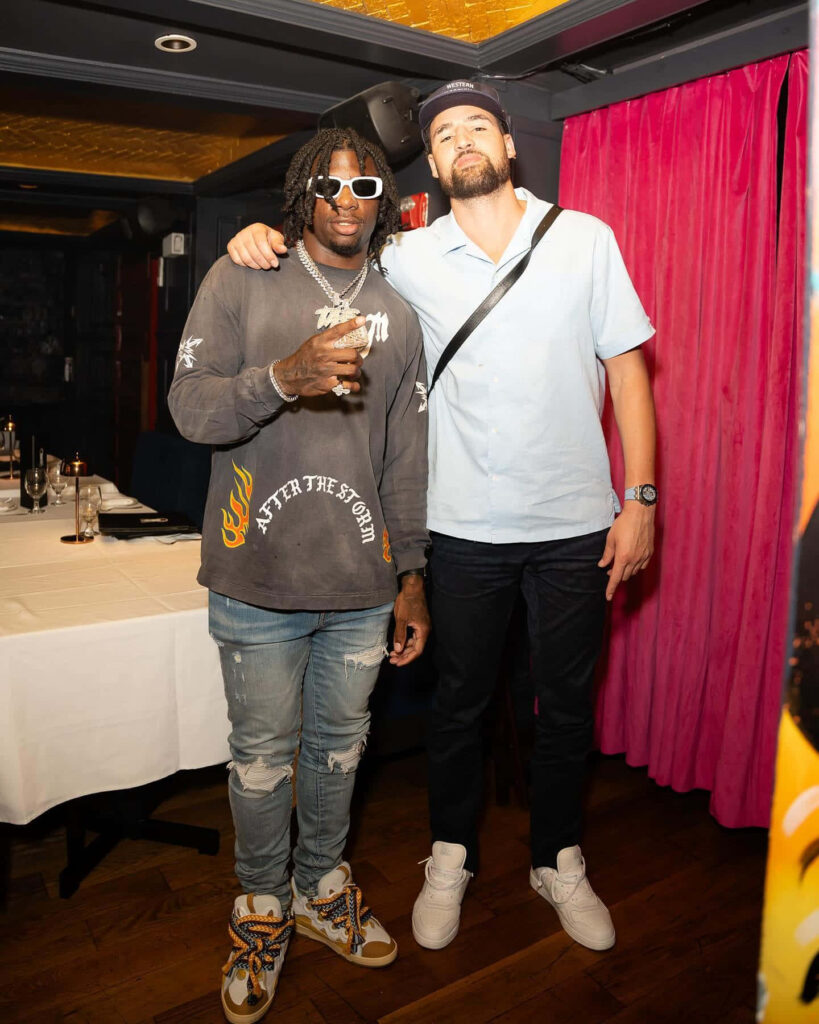 Deonte Banks in graphic sweatshirt and ripped jeans with friend in stylish indoor setting vibrant colors Wallpaper
