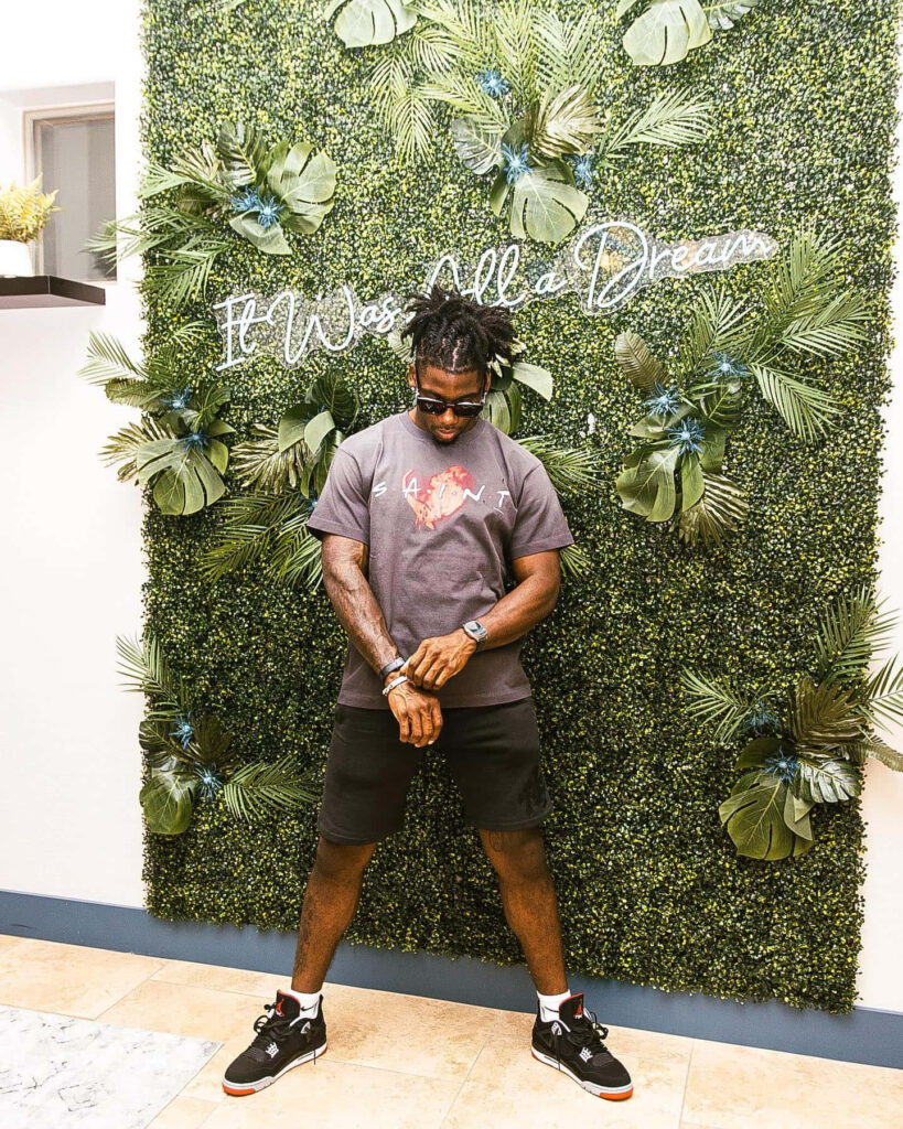 Deonte Banks poses confidently by green foliage and neon It Was All a Dream sign urban fashion wallpaper