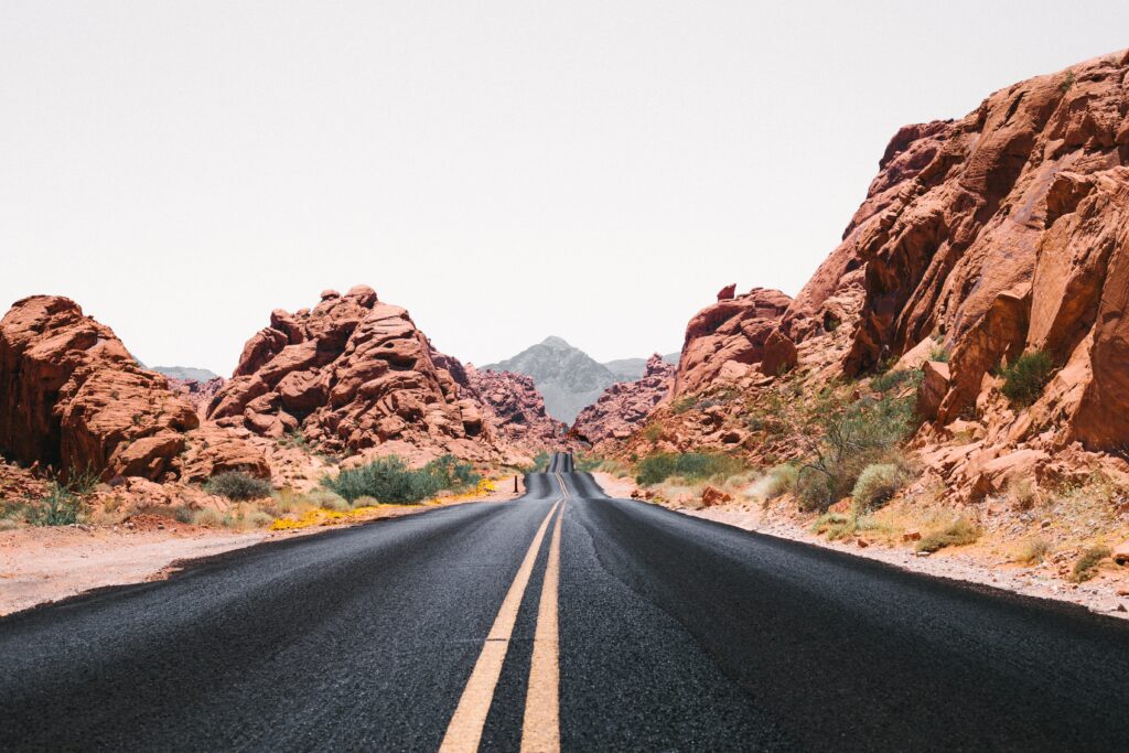 Straight road with yellow lines through red desert rocks and towering formations under pale sky Wallpaper