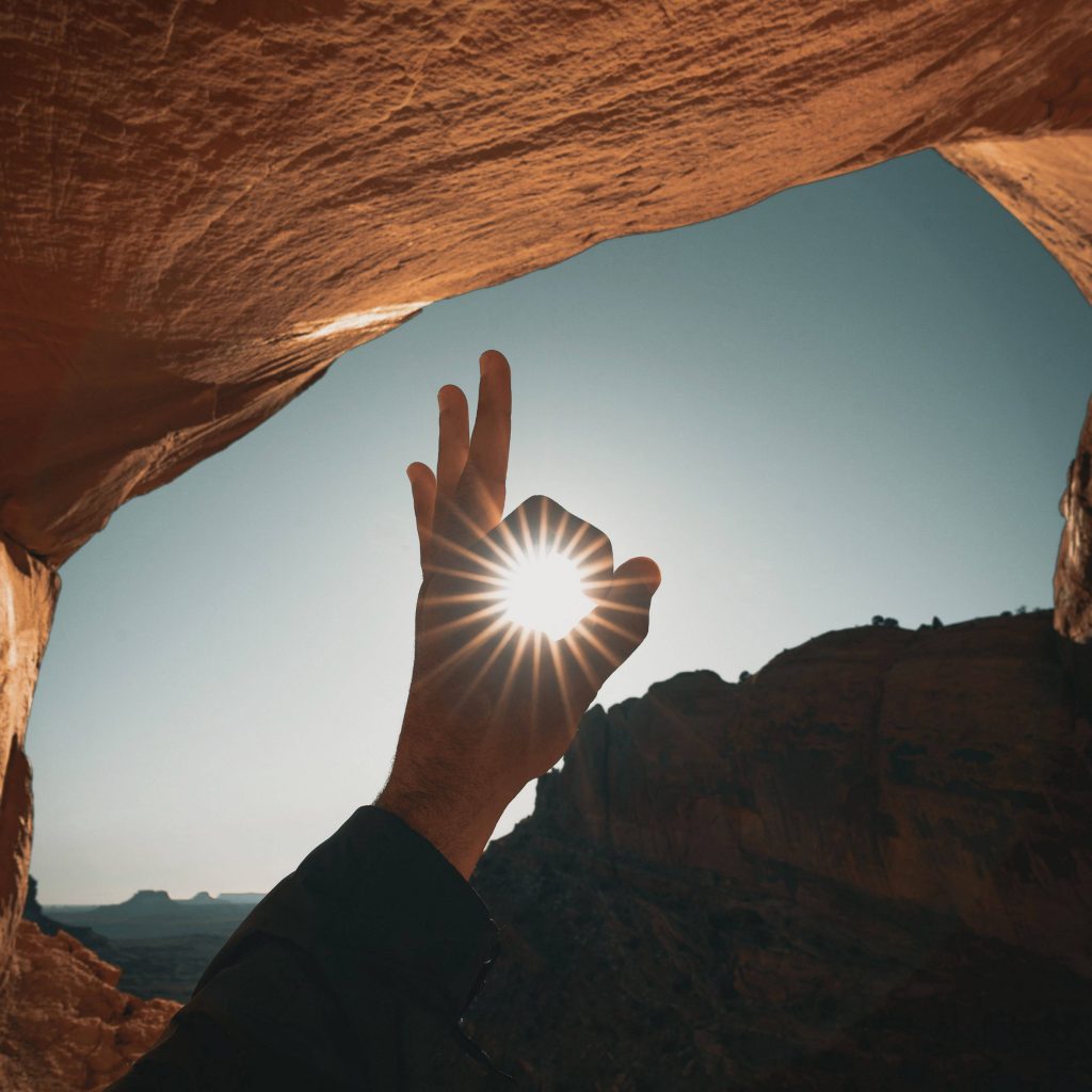 Hand framing sun with OK gesture inside sandstone arch glowing golden desert light wallpaper