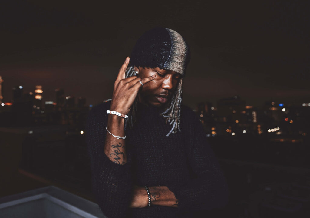 Destroy Lonely rapper on rooftop at night with city lights wearing beanie and dark sweater Wallpaper
