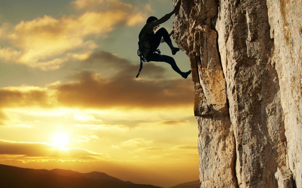 Man climbing rugged rock face at sunset golden sky adventure nature wallpaper background