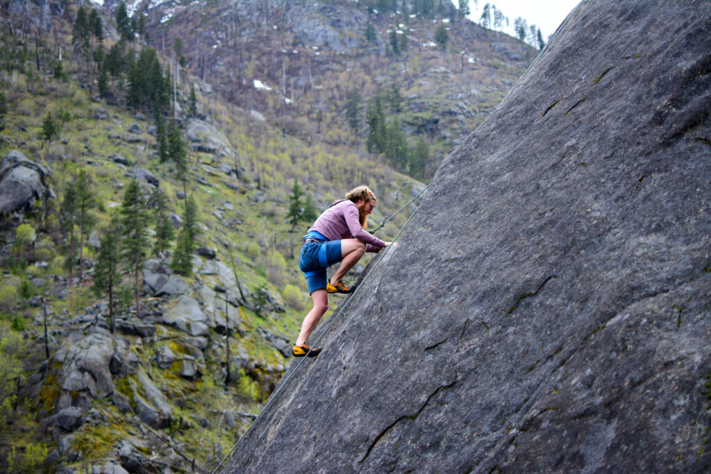 Determined rock climber ascending steep cliff with rugged mountains and pine trees background Wallpaper