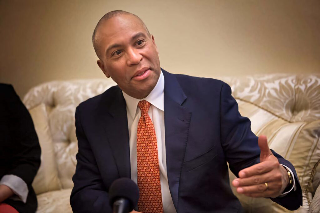 Deval Patrick speaking in media interview seated on cream sofa wearing navy suit and orange tie Wallpaper