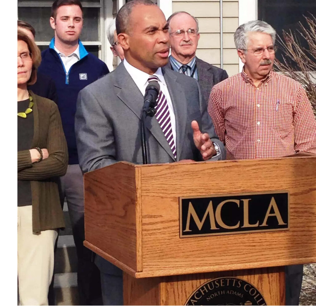 Deval Patrick speaking at MCLA podium outdoors in gray suit with audience and academic buildings Wallpaper
