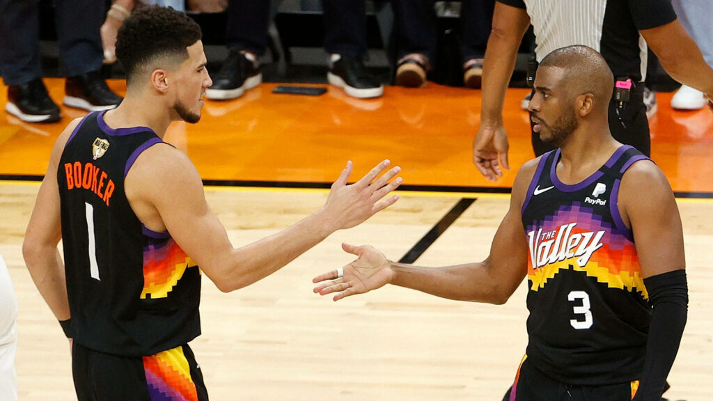 Devin Booker and Chris Paul in Phoenix Suns jerseys high-five on court with sunset background Wallpaper