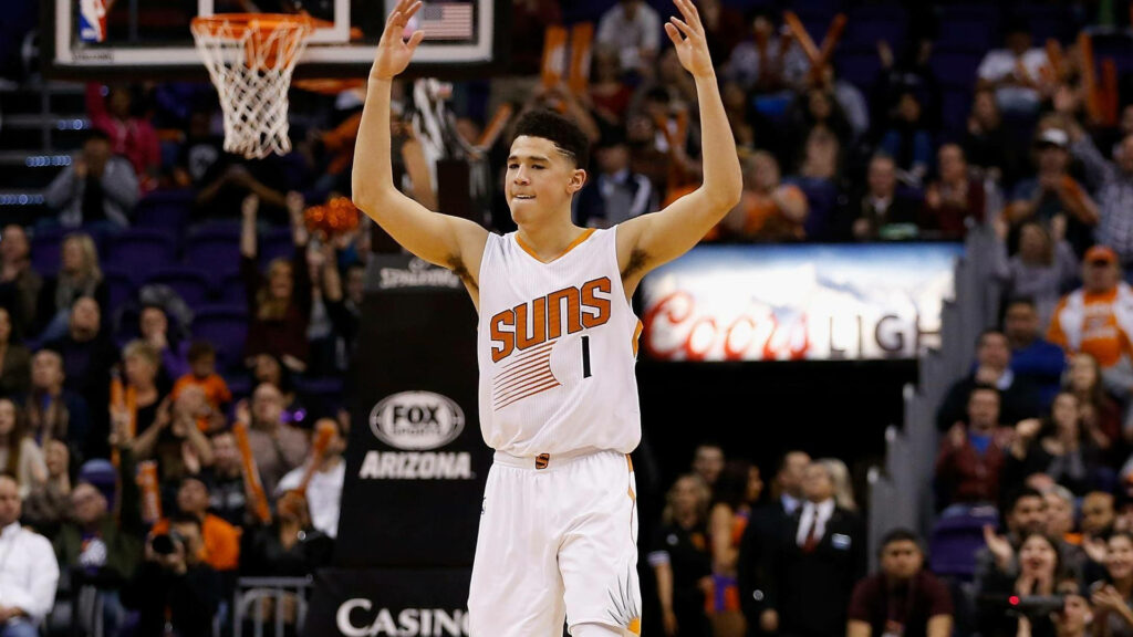 Devin Booker celebrates on court in Phoenix Suns uniform with fans and arena lights background Wallpaper