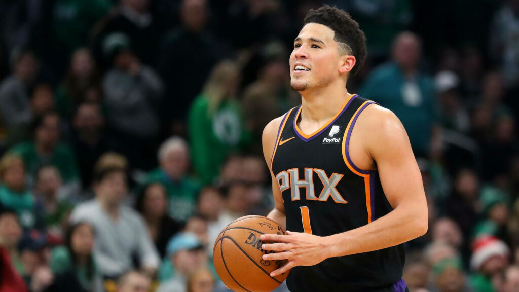 Devin Booker in black Suns jersey holding basketball on court with vibrant crowd and stadium lights Wallpaper