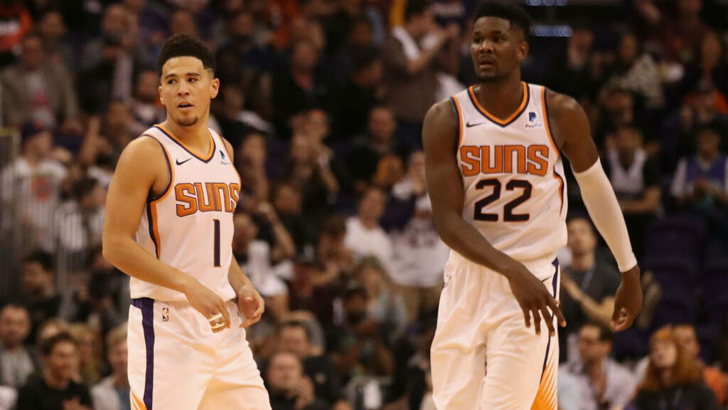 Devin Booker and Deandre Ayton in white Phoenix Suns jerseys on court with vibrant crowd background Wallpaper