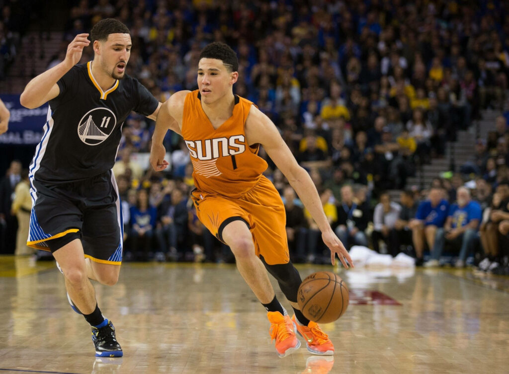 Devin Booker dribbling past Klay Thompson in intense NBA game vibrant Suns and Warriors jerseys Wallpaper