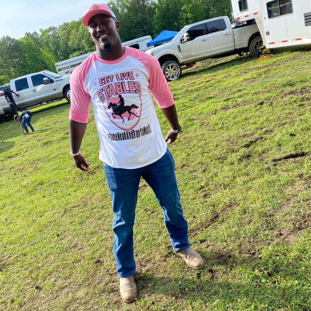 Devin White smiling outdoors in pink Get Live Stables shirt and cap with trucks and field background Wallpaper