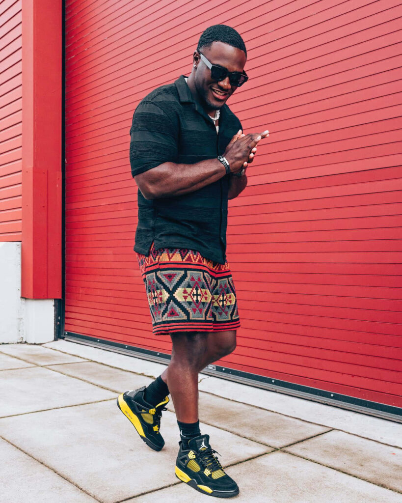 Devin White smiling texting in black shirt patterned shorts red wall vibrant contemporary wallpaper background