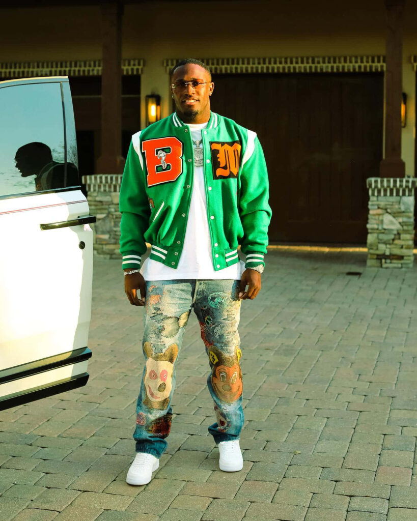 Devin White confident in green varsity jacket and embroidered jeans with colorful patches outdoors Wallpaper