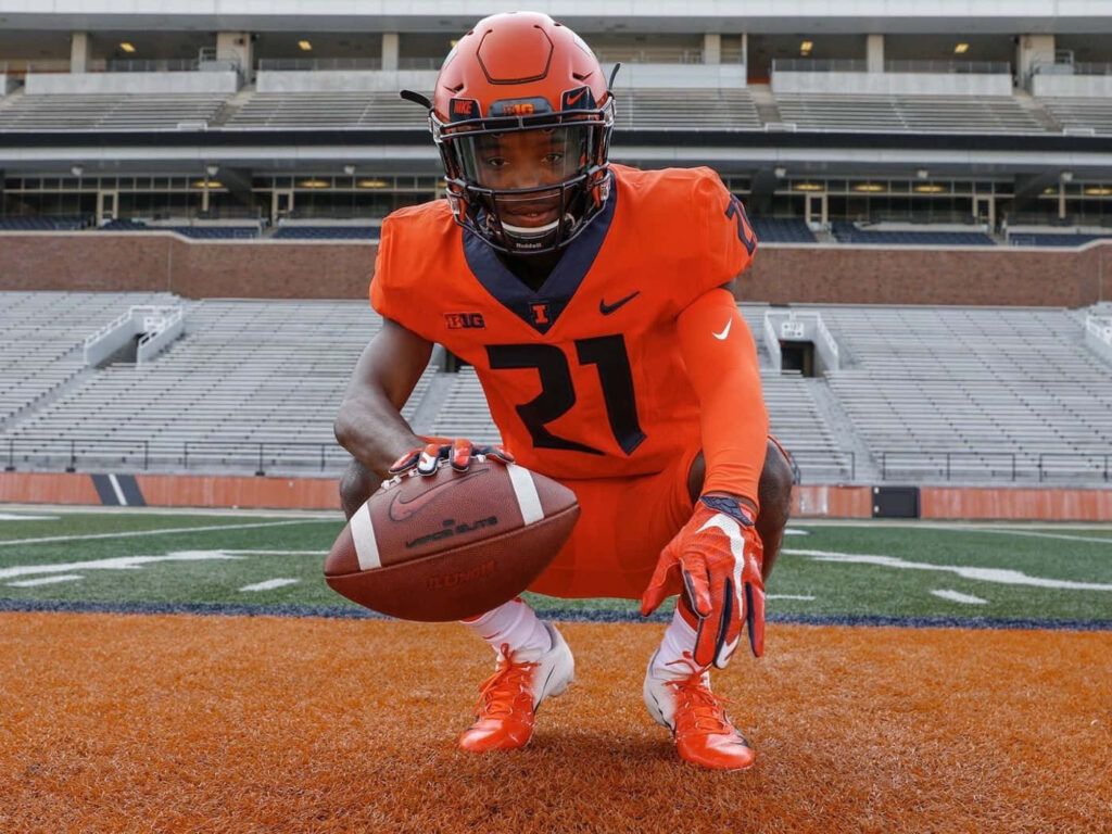 Devon Witherspoon in orange Illinois football uniform crouched ready with ball on stadium field background Wallpaper